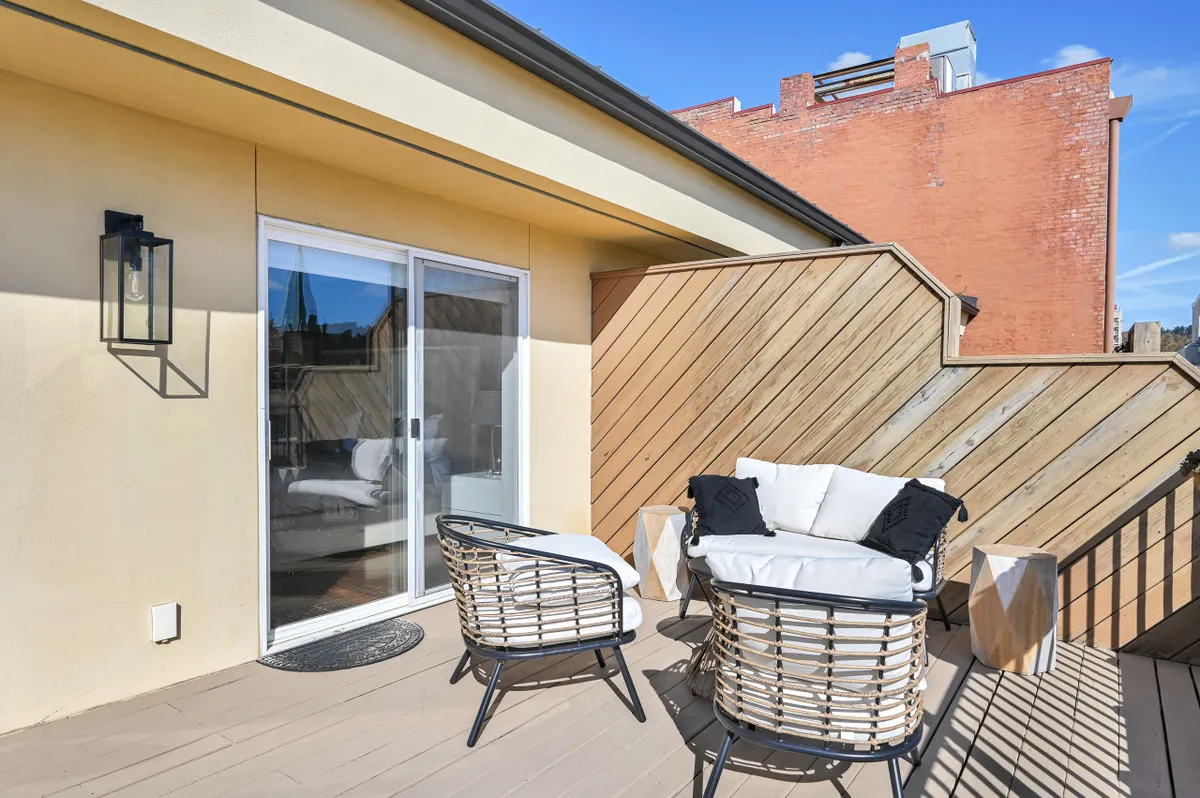 This exterior image features a cozy outdoor seating area bordered by a wooden privacy wall and accessed via a sleek sliding glass door. The space is bathed in warm light, suggesting both morning coffee and cozy evening gatherings. The cityscape is partially visible, hinting at the urban setting while emphasizing privacy and comfort.
