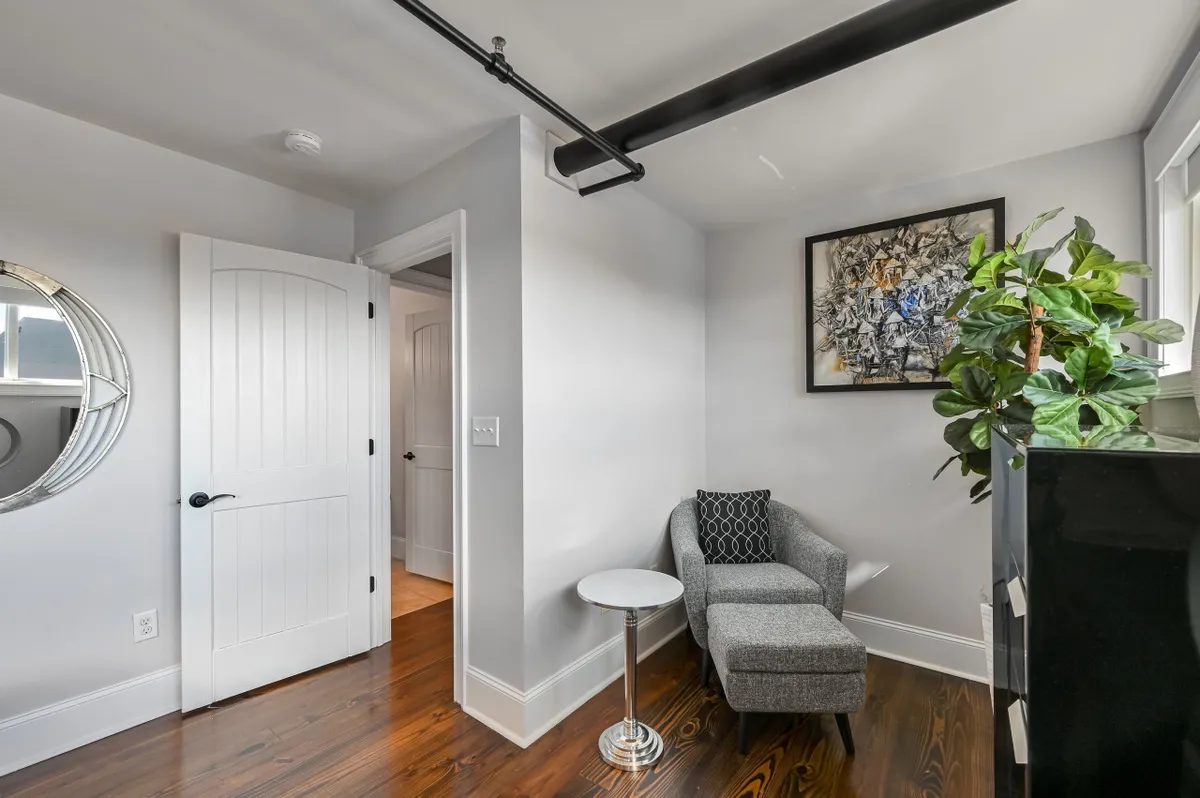 A calm, cozy sitting area with a grey chaise lounge, round side table, abstract wall art, and a green plant. Morning light streams into the space, inviting relaxation and quiet conversation.