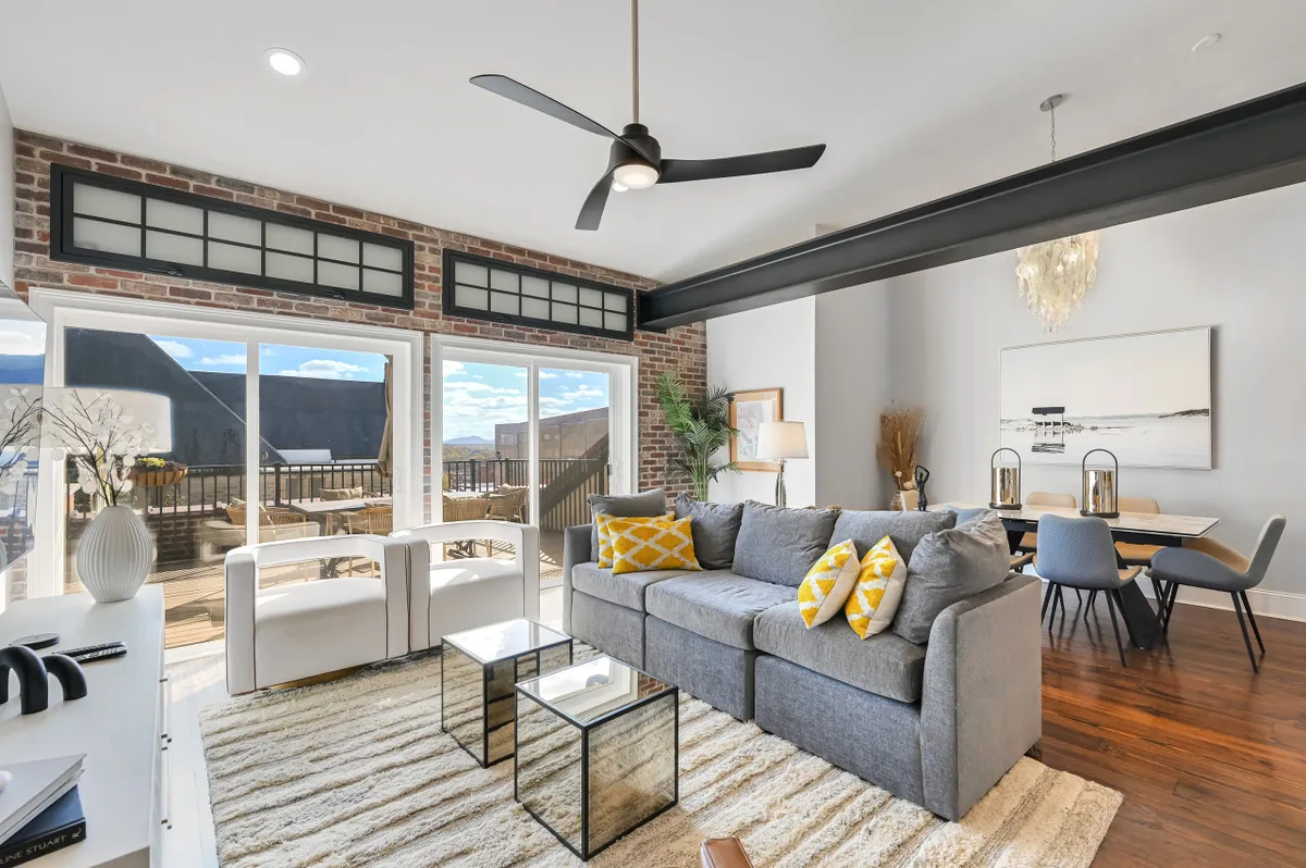 This image features a modern living space with a sofa adorned with yellow pillows, a glass coffee table, and large windows letting in plenty of natural light. The windows frame a view of the city, and the dining area is visible in the background, contributing to an open and inviting atmosphere.