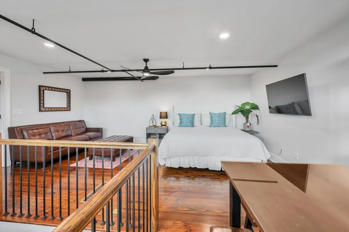 A versatile loft area with a bed adorned with decorative pillows, a modern sofa, a dining or work table, a large wall-mounted TV, and a decorative plant. The space is bright, open, and adaptable, suited for relaxing, working, or socializing.