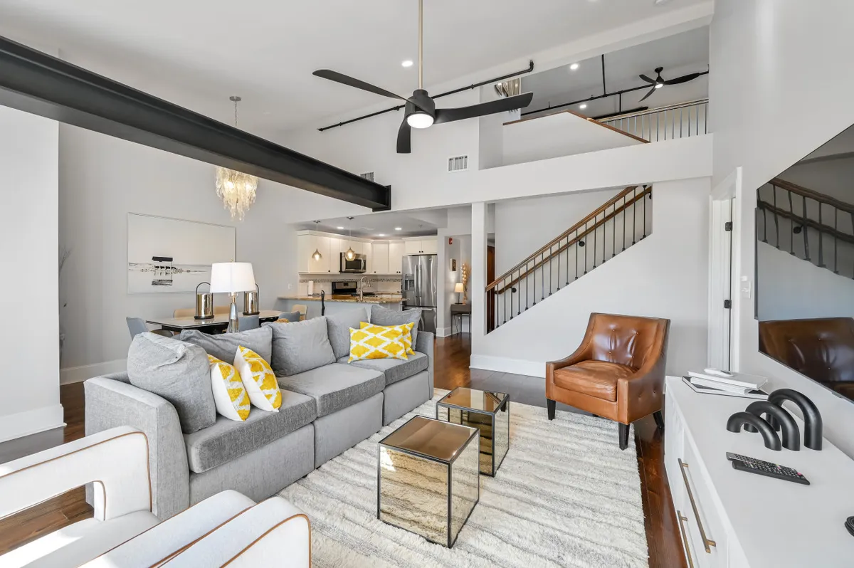 A spacious, modern living area featuring a sofa with colorful cushions, a wooden staircase, a contemporary ceiling fan, a chandelier, and a glass coffee table. The room feels airy and open, with design elements that hint at a connection to upper floors and possibly the terrace.