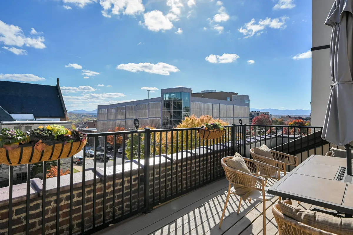 This exterior image presents a long terrace with a balcony railing, vibrant planters filled with flowers, comfortable outdoor seating, and a table. The terrace overlooks Asheville's downtown skyline, which glows with autumnal colors and city lights. The mood is relaxed, with people enjoying coffee, reading, or entertaining guests while taking in expansive city views.
