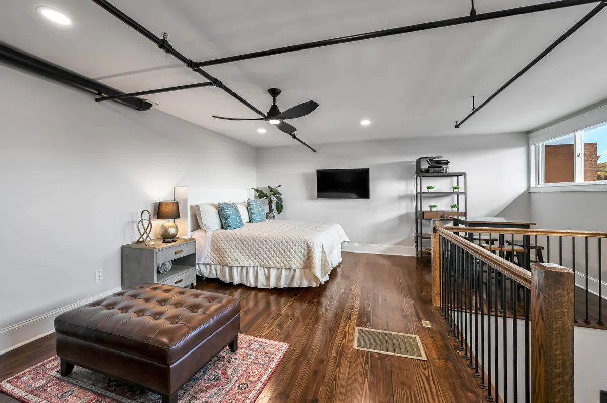 A modern bedroom featuring a bed with decorative pillows, a leather ottoman, a bedside lamp, a wall-mounted TV, a decorative plant, and a bookshelf. The space feels cozy, contemporary, and designed for restful moments together.