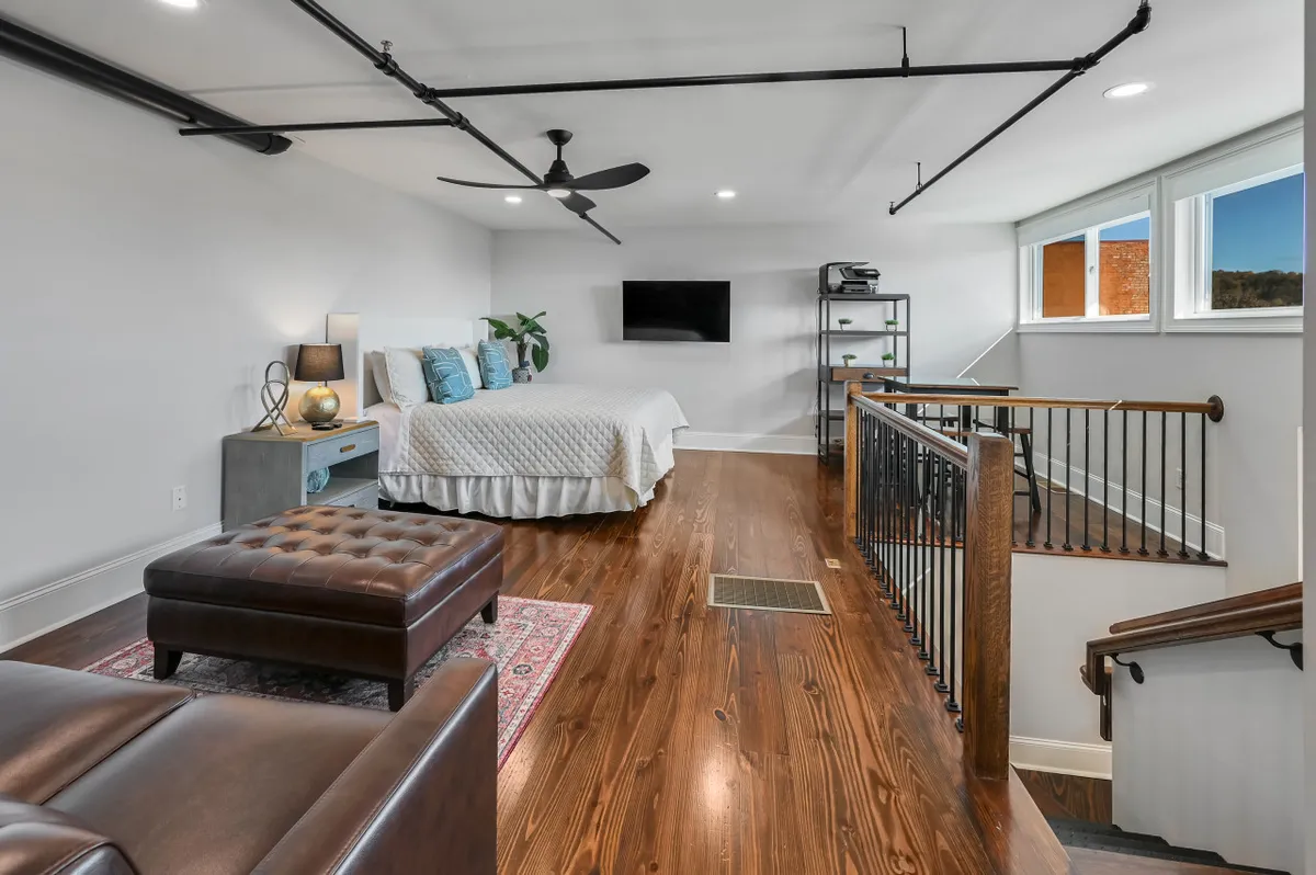 A stylish bedroom with decorative pillows on the bed, a brown ottoman, wall-mounted TV, and open windows letting in abundant natural light. The windows provide views of the outside, suggesting a connection to the surrounding cityscape and terrace area.