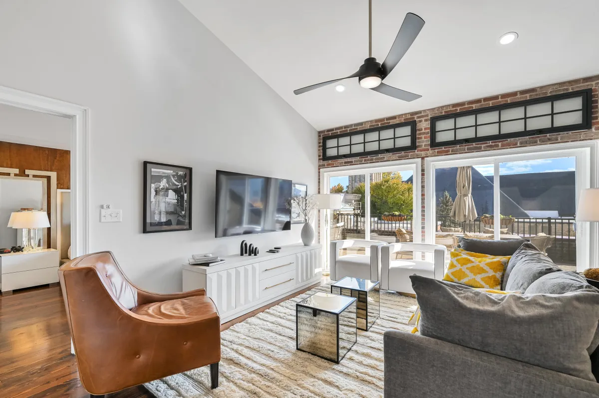 This image showcases a sunlit living area with a plush brown leather chair, a sleek white TV console, and expansive windows offering south-facing city views. The space is accented by a pop of yellow from an accent pillow, blending contemporary comfort with the historic character of the building. The inviting setup hints at relaxation, conversation, and taking in the city skyline.