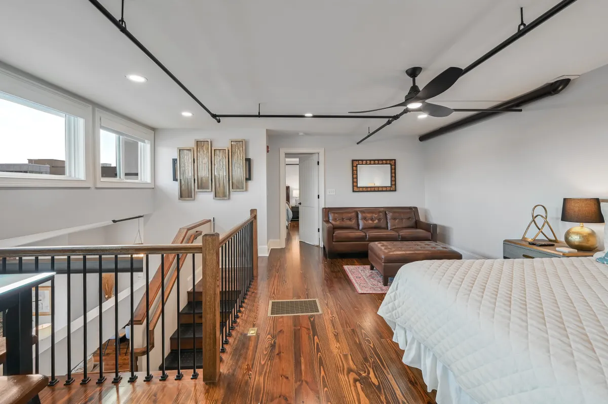 A view of a bright, comfortable interior featuring a wooden staircase, leather sofa, decorative wall art, and a bed with a quilted cover. Natural light pours in, illuminating both the seating and sleeping areas, and the space artfully balances comfort with modern aesthetics.